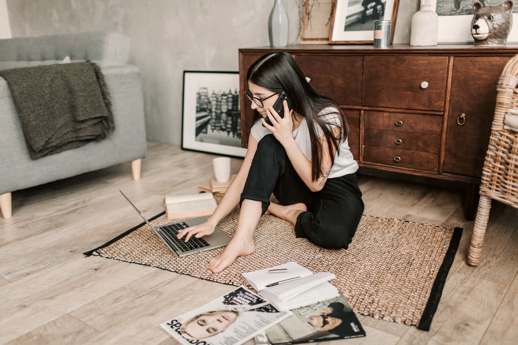 Mulher sentada no chão da sala falando ao telefone enquanto usa o notebook, cercada de papéis